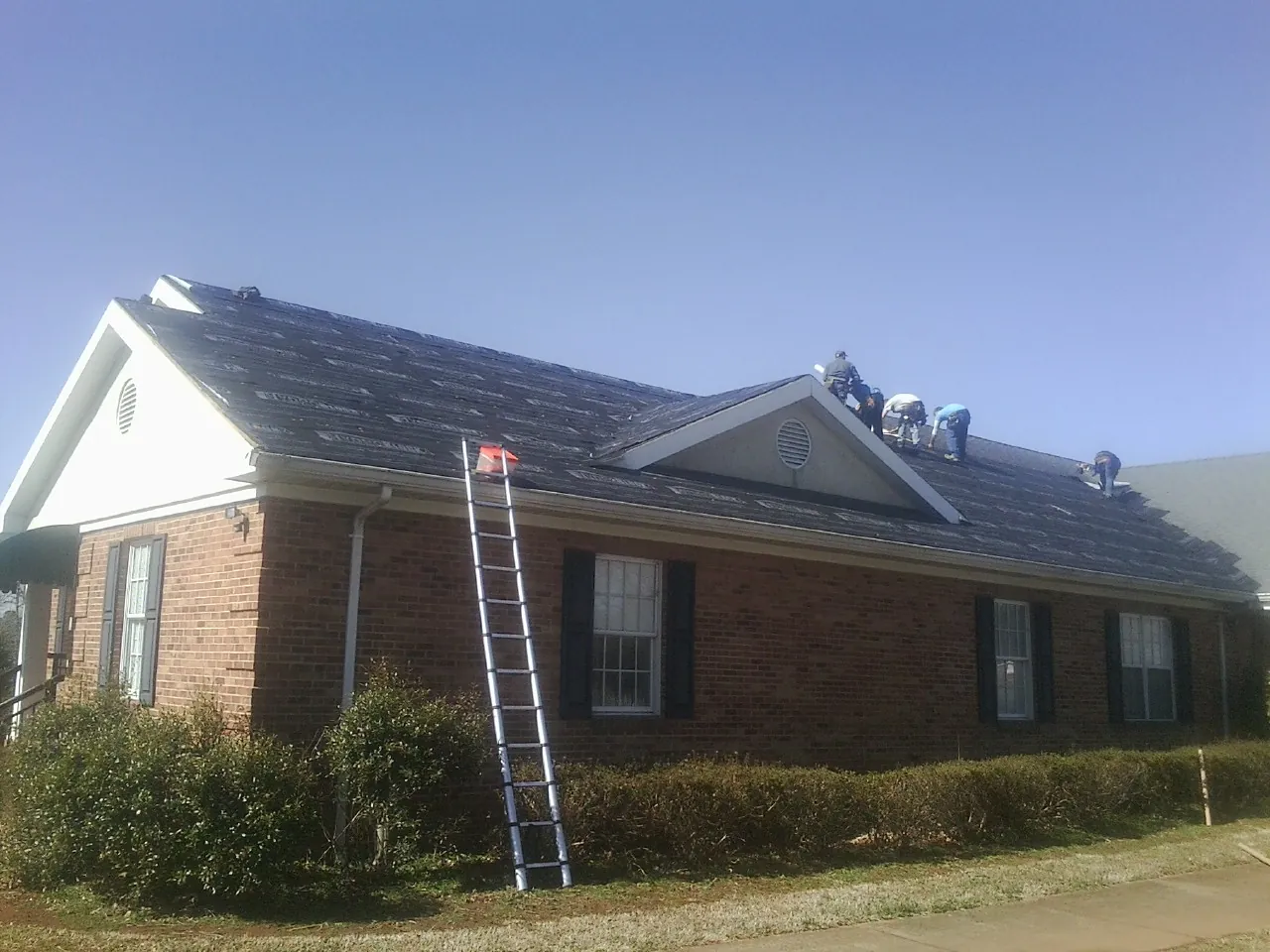 Crew tearing shingles off a brick ranch roof with a ladder set — Metal Roof Repair work by our crew in Ramah
